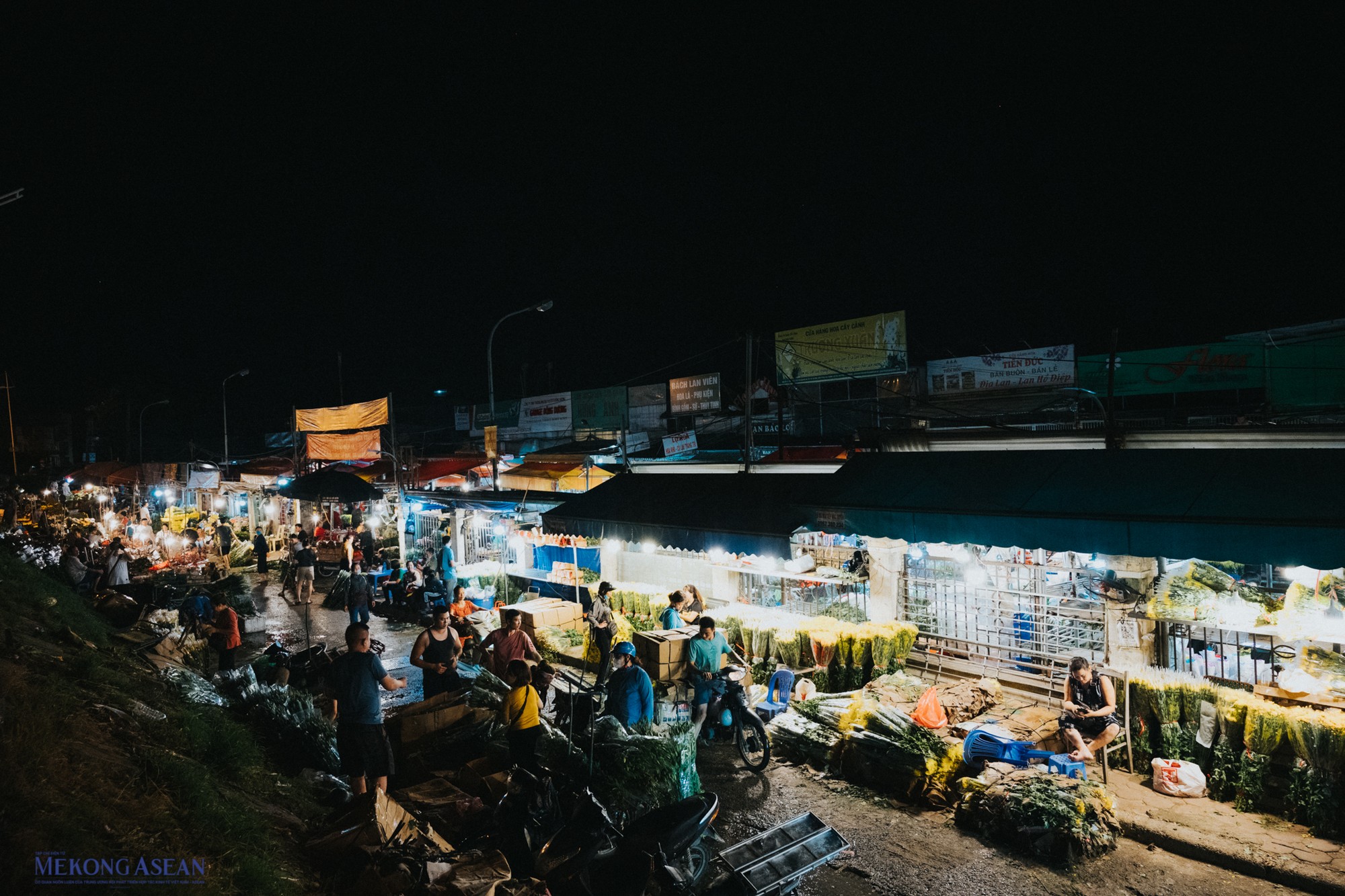 Khung cảnh từ tr&ecirc;n đường đ&ecirc; s&ocirc;ng Hồng nh&igrave;n xuống, ph&iacute;a ngo&agrave;i chợ d&agrave;i khoảng 500m với những h&agrave;ng hoa li&ecirc;n tiếp tạo th&agrave;nh một d&atilde;y đường s&aacute;ng lung linh suốt đ&ecirc;m.